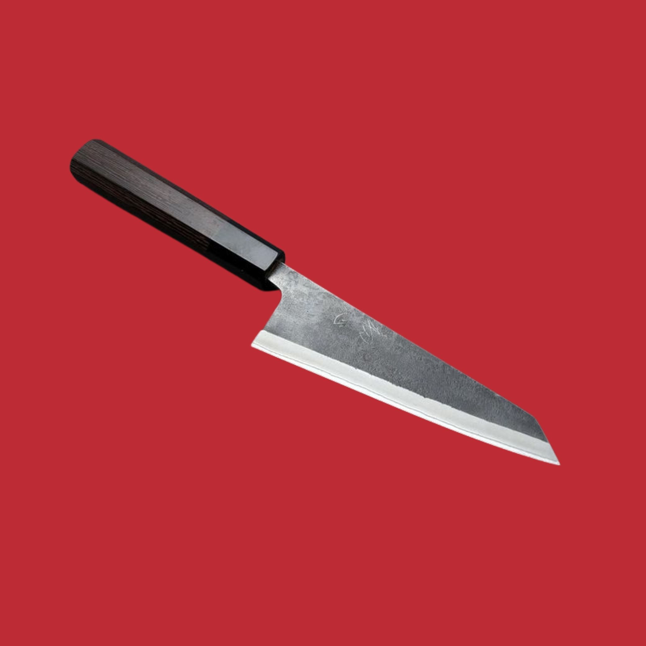 An artisanal kitchen knife with Japanese inscriptions on its matte blade and a wooden, dark handle sits diagonally against a red background.