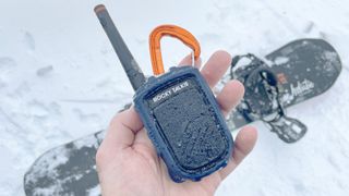 Close-up of a Rocky Talkie Mountain Radio walkie-talkie in a user's hand with a snowboard and snow in the background