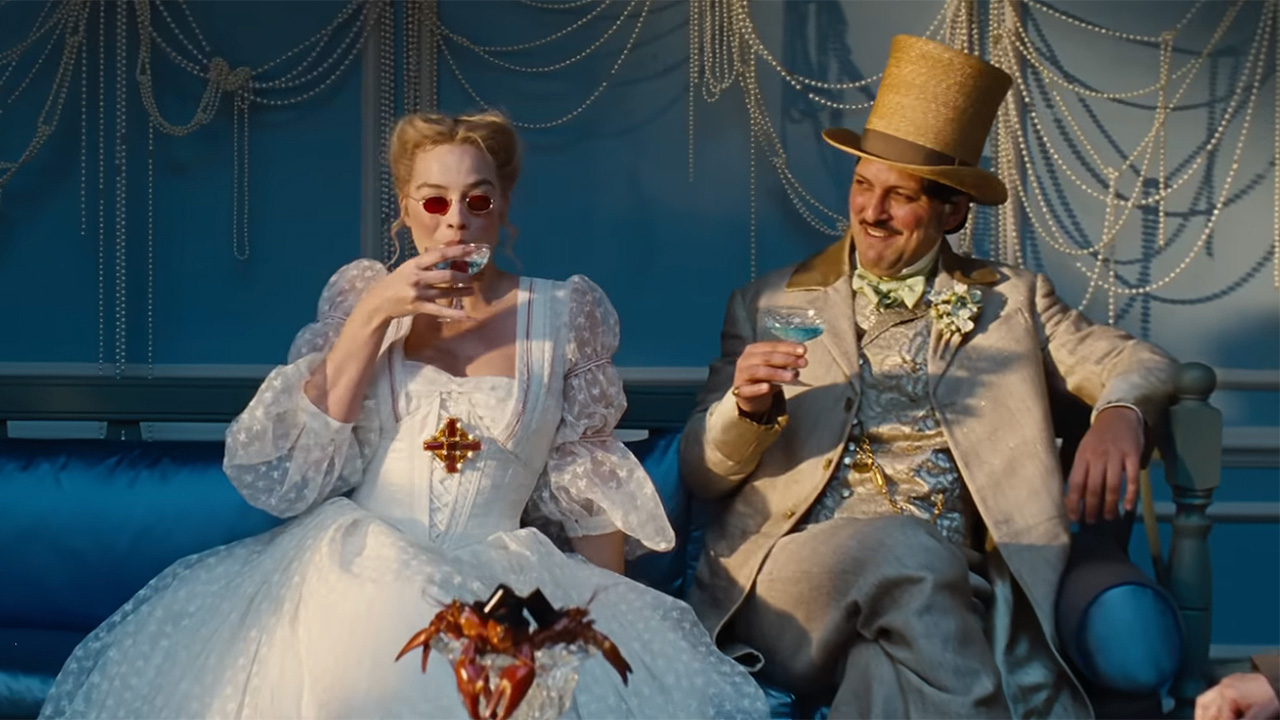 Margot Robbie sips a cocktail next to a gentleman on a blue coach in a white dress and wild sunglasses in Wuthering Heights. 