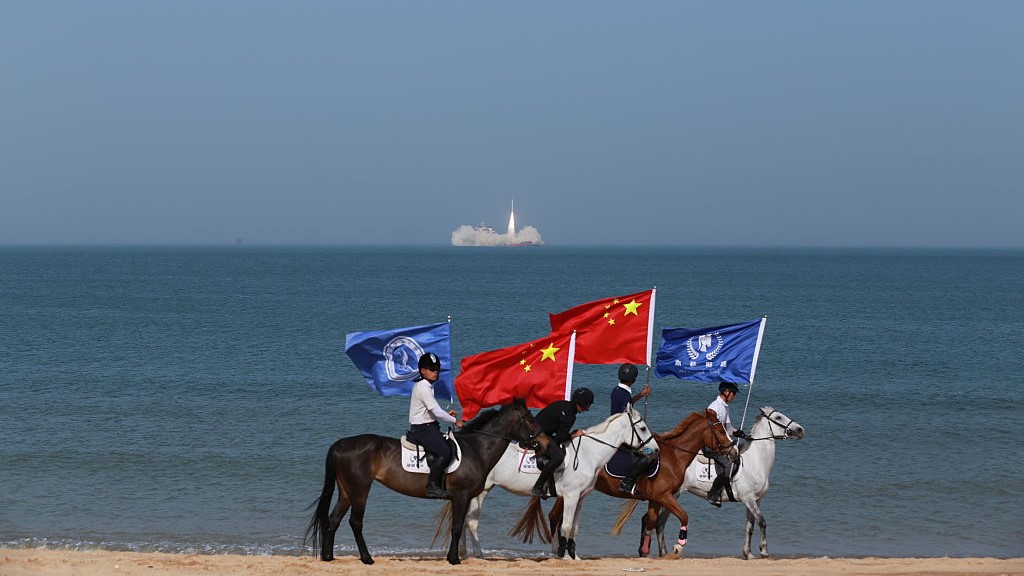 On a beach next to the sea, a man in a white shirt holding a blue flag on a brown horse looking away from the sea behind a man in a black shirt holding a red flag on a white horse looking toward the sea behind a man in a black shirt with a red flag on a brown horse looking toward the sea behind a man in a white shirt holding a blue flag on a white horse looking ahead to the right. On the horizon of the sea, a small white rocket rises from a plume of smoke. 