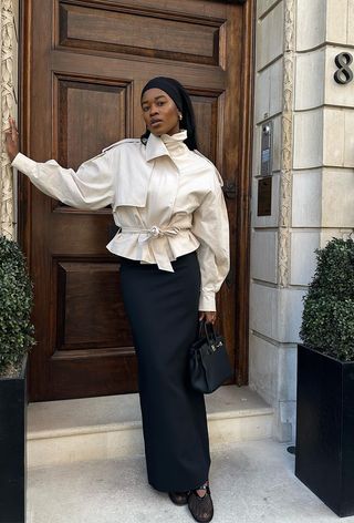 best cropped trench coat for women shown on a woman standing in a doorway wearing a light tan cropped trench coat with a black maxi skirt, black mesh ballet flats, and a black handbag