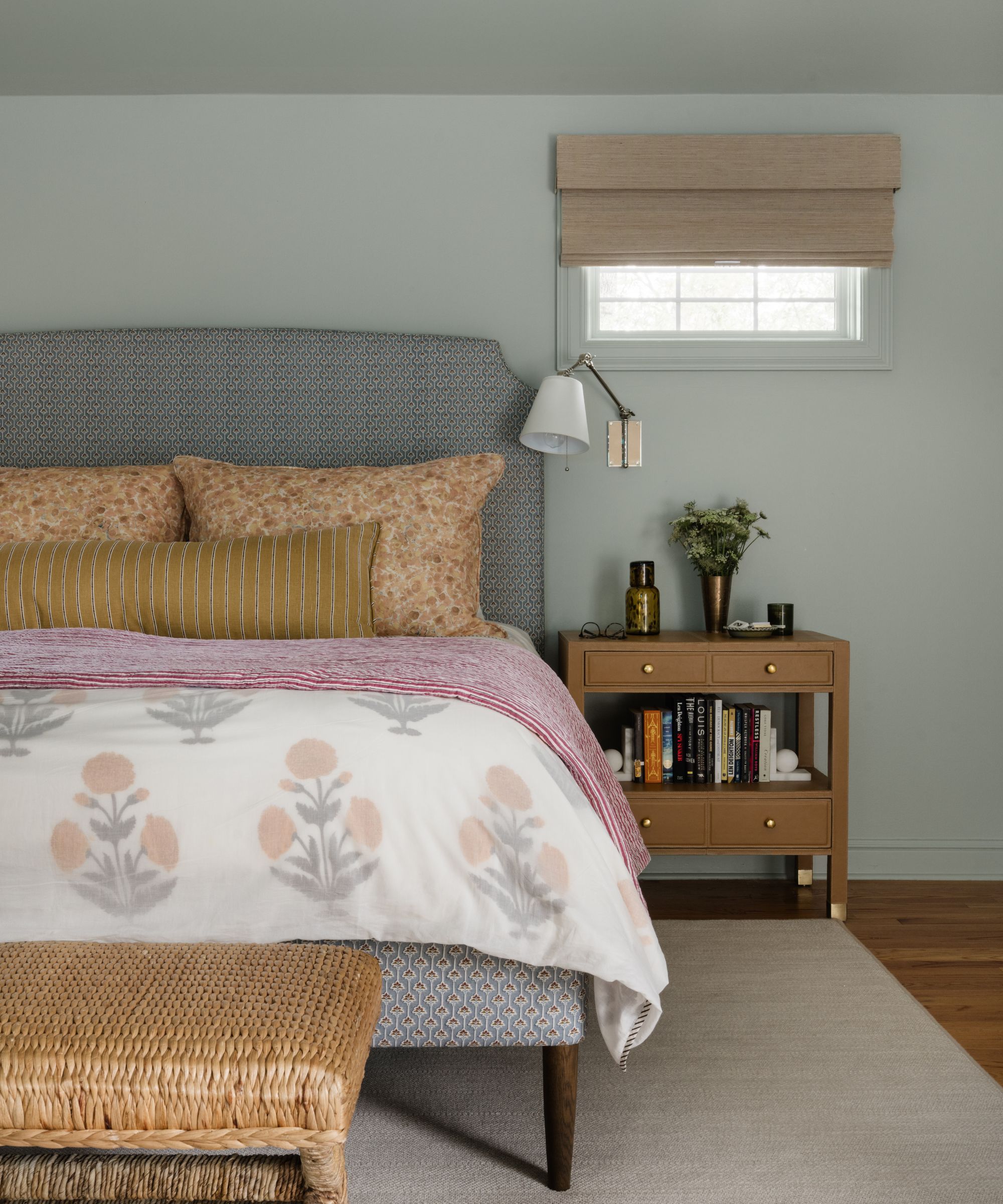 A calming bedroom with gray-green color-drenched walls, a gray bed with orange and pink bedding, and a brown nightstand.