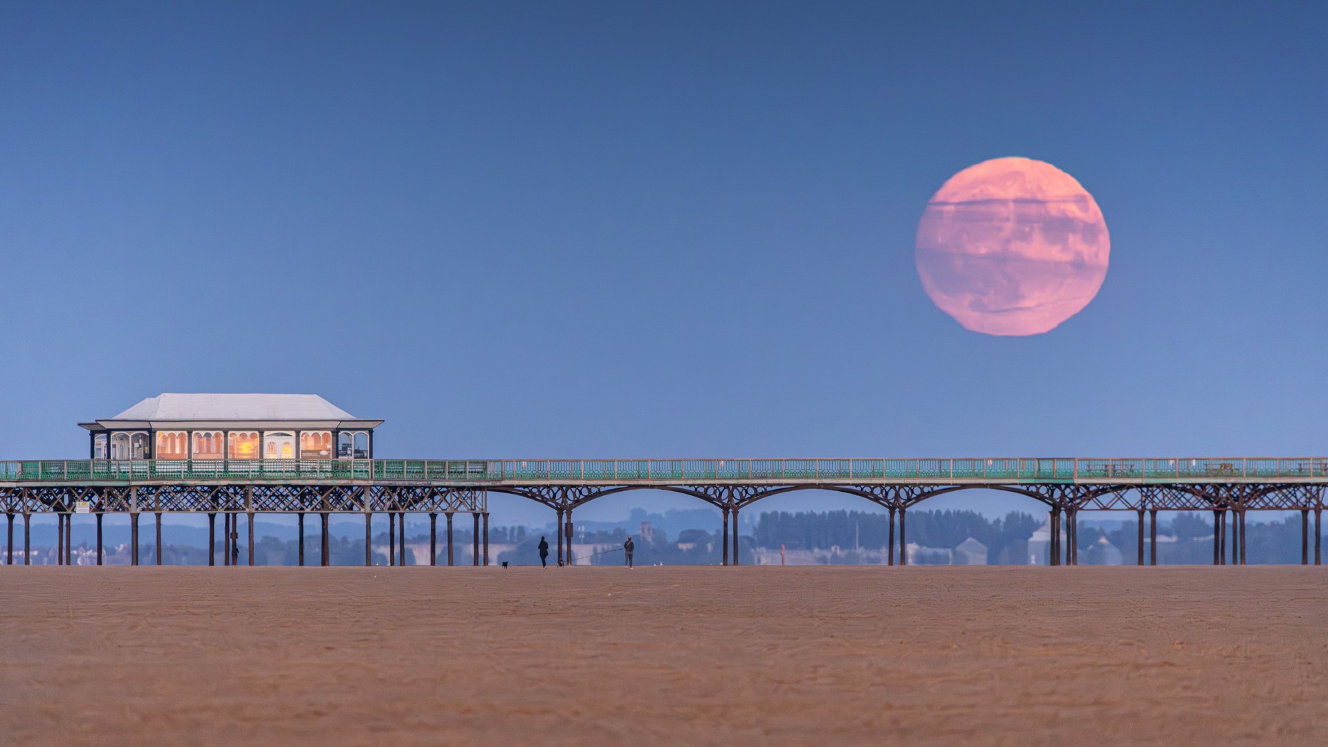An orange full moon is pictured rising over a sandy beach, on which sits a large pier. The sky is turning a dark blue, and sunlight can be seen reflected in the windows of a building on the pier.