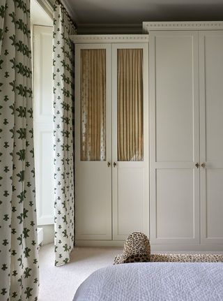 bedroom with built-in closet with glass-fronted cabinets with ruffled curtains behind, patterned curtains over window and leopard print bench at the end of the bed