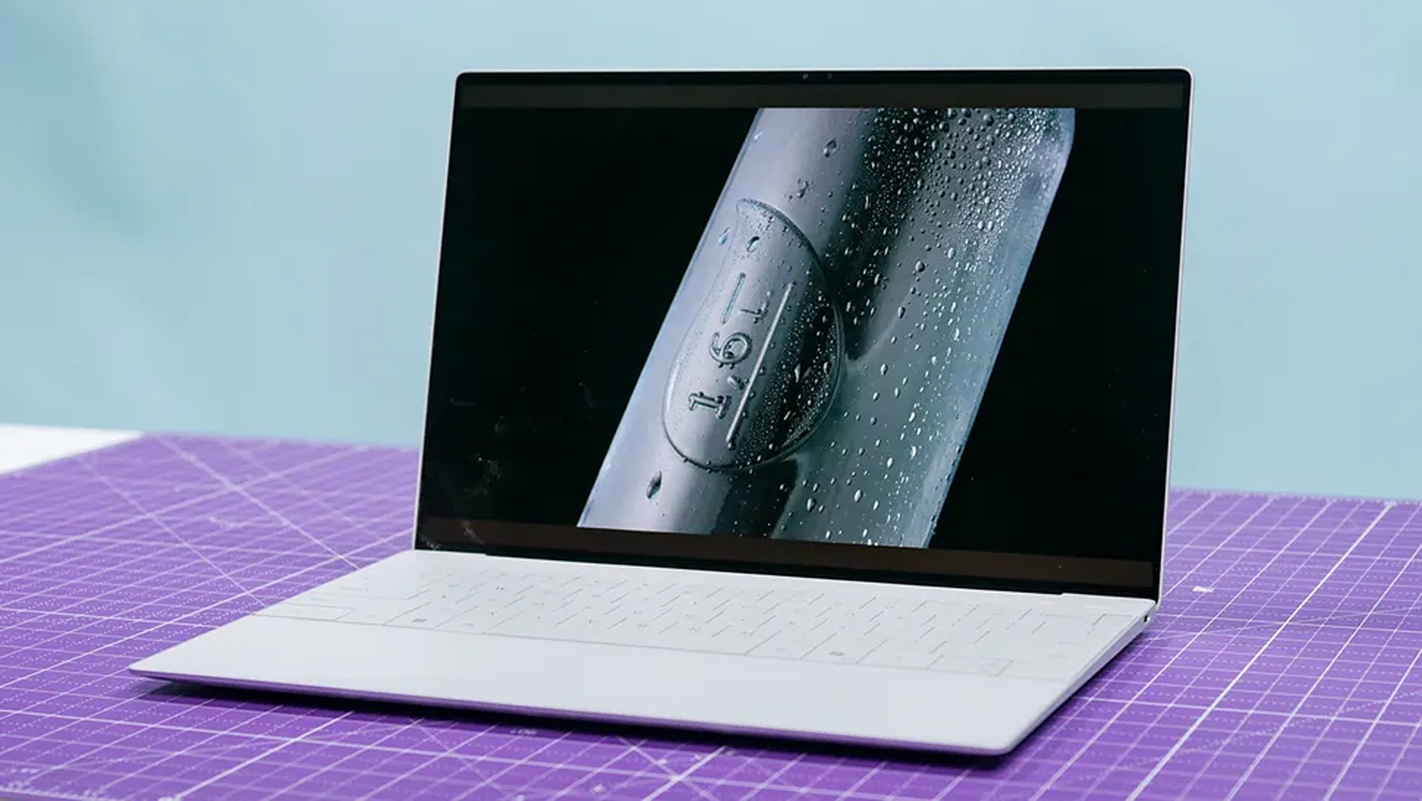 A sleek, white laptop on a purple grid-patterned surface. Its screen displays a close-up of a metallic object with water droplets, creating a modern tech vibe.