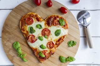 A heart shaped pizza on a round wooden board with a pizza cutter, basil leaves and cherry tomatoes.
