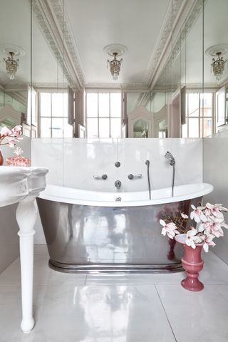 A luxury bathroom with reflective roll top freestanding bathtub and mirrored wall and white wall and floor tiles and vanity