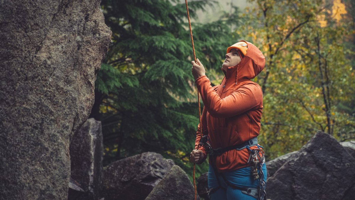 A rock climber belays at the bottom of a cliff surrounded by trees with autumn coloured leafs 