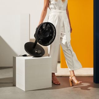 Duux Whisper Flex Smart Standing Fan being picked up by woman in white jumpsuit in yellow and white room