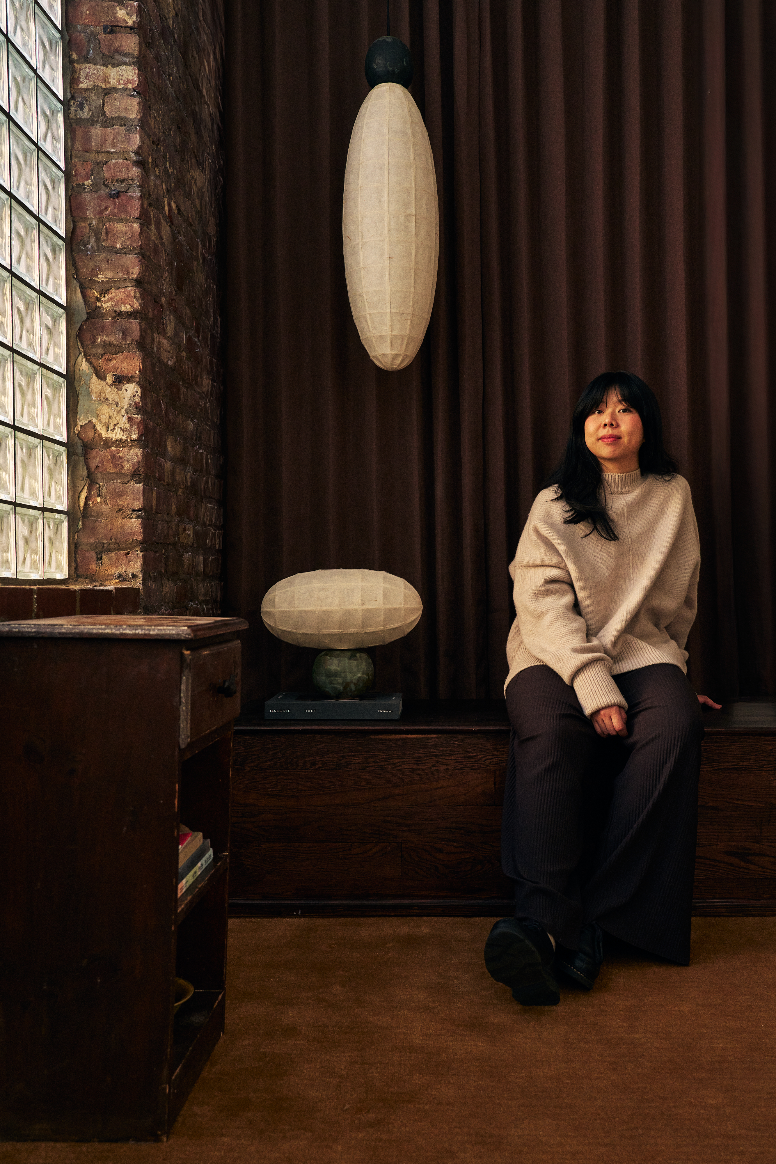 A young woman in a loose-fitting gray jumper and dark trousers sits in a design showroom painted a chocolatey brown, with glass block windows, next to some paper lamp creations.