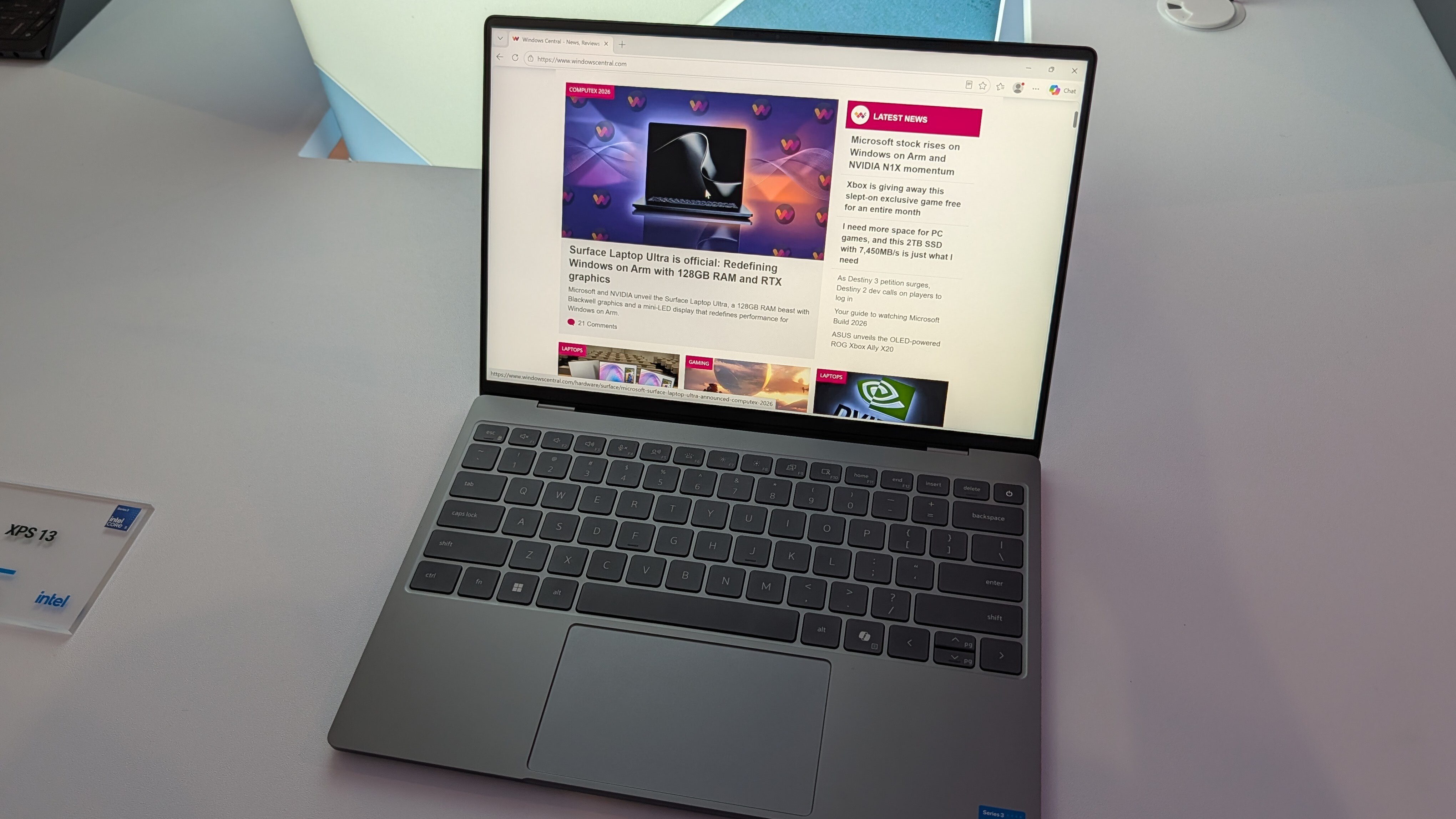 An open XPS 13 laptop displaying a webpage with tech articles on a light-colored surface, next to a card labeled "XPS 13" with the Intel logo.