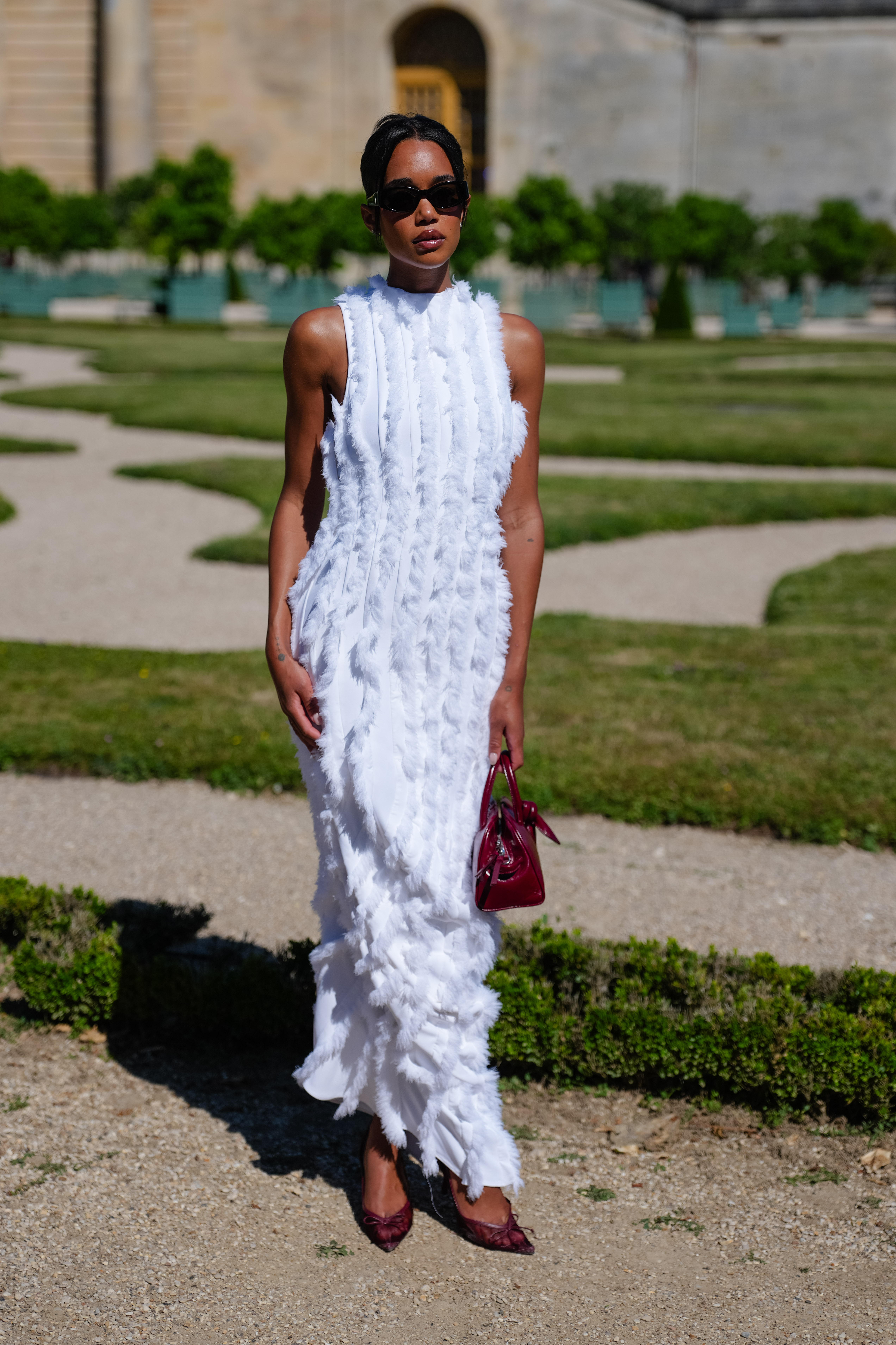 VERSAILLES, FRANCE - JUNE 29: Laura Harrier wears a sleeveless white floor-length dress with textured vertical ruffled stripes and a high neckline, styled with dark burgundy pointed-toe heels and a glossy burgundy croc-embossed leather handbag. Hair is pulled back in a sleek low bun and makeup includes bold brows and a matte lip. Black rectangular sunglasses complete the look, outside Jacquemus, during the Paris Fashion Week - Menswear Spring/Summer 2026, on June 29, 2025 in Versailles, France.