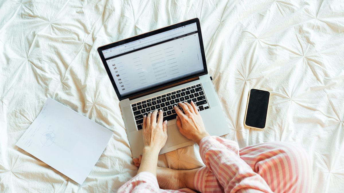 A laptop, phone and notebook lying on a mattress, a pair of hands is typing on the laptop