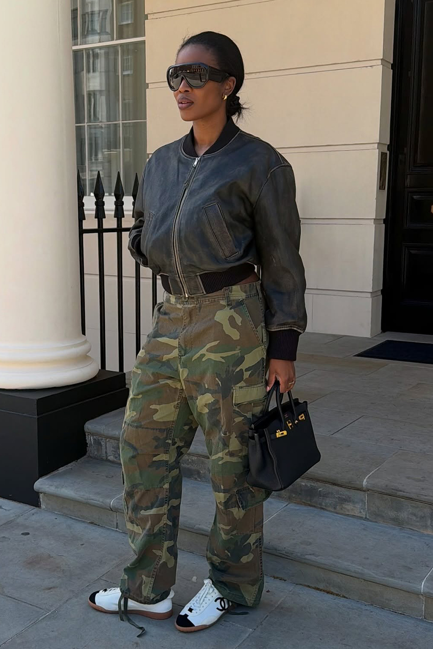 A woman wearing a brown-black leather bomber jacket, camouflage cargo pants, white and black Chanel sneakers, and a Hermes black leather bag