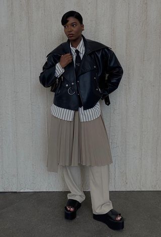 A woman wearing a black leather moto jacket with a striped button-down shirt, a black tie, a tan pleated skirt layered over tan trousers, and black platform sandals.