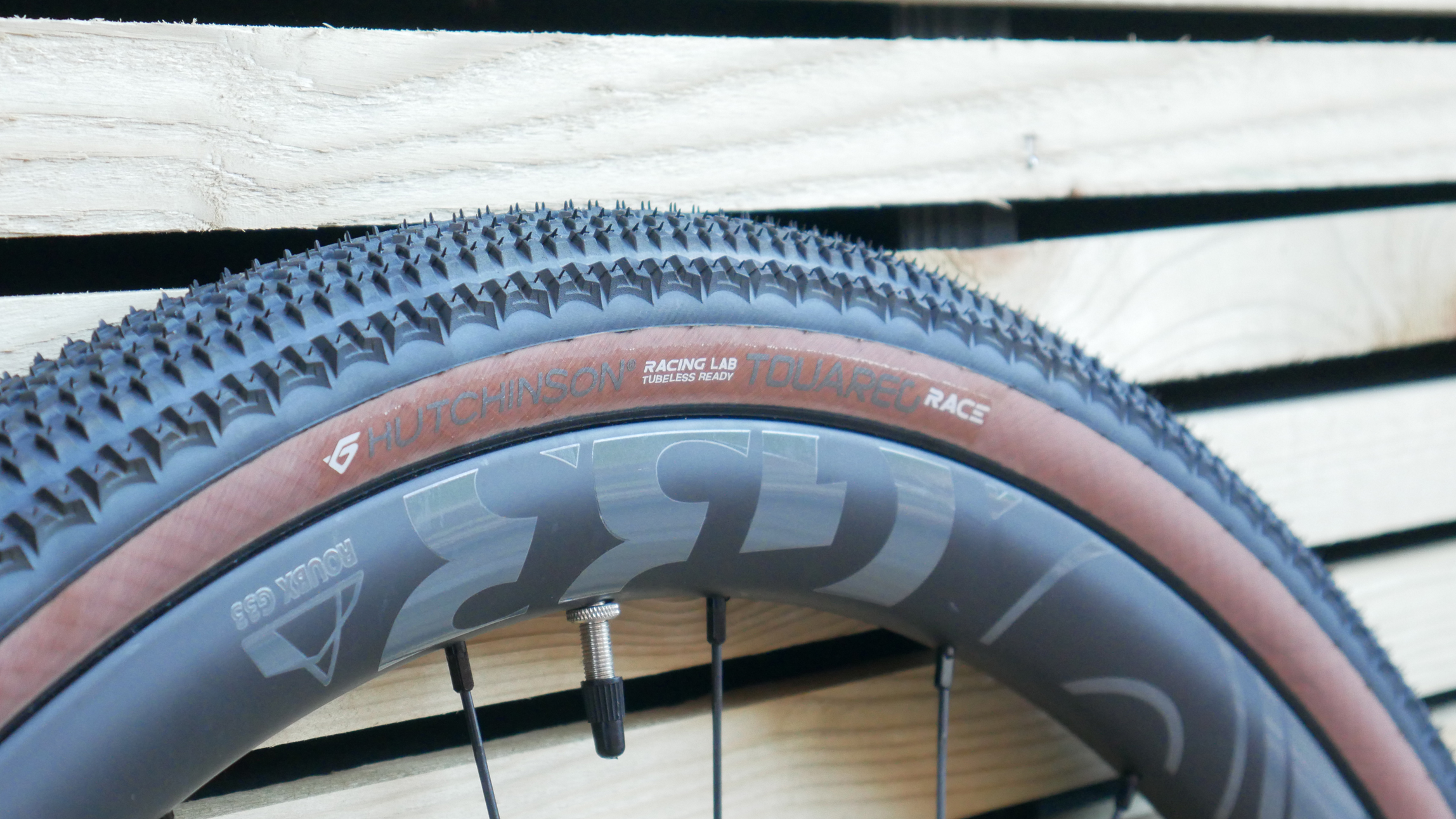 A close-up of the Hutchinson Touareg Race 45mm tread profile and shape specifically the Hutchinson Touareg heatpatch logo branded on the dark tan wall sidewalls, with wooden cladding in the background