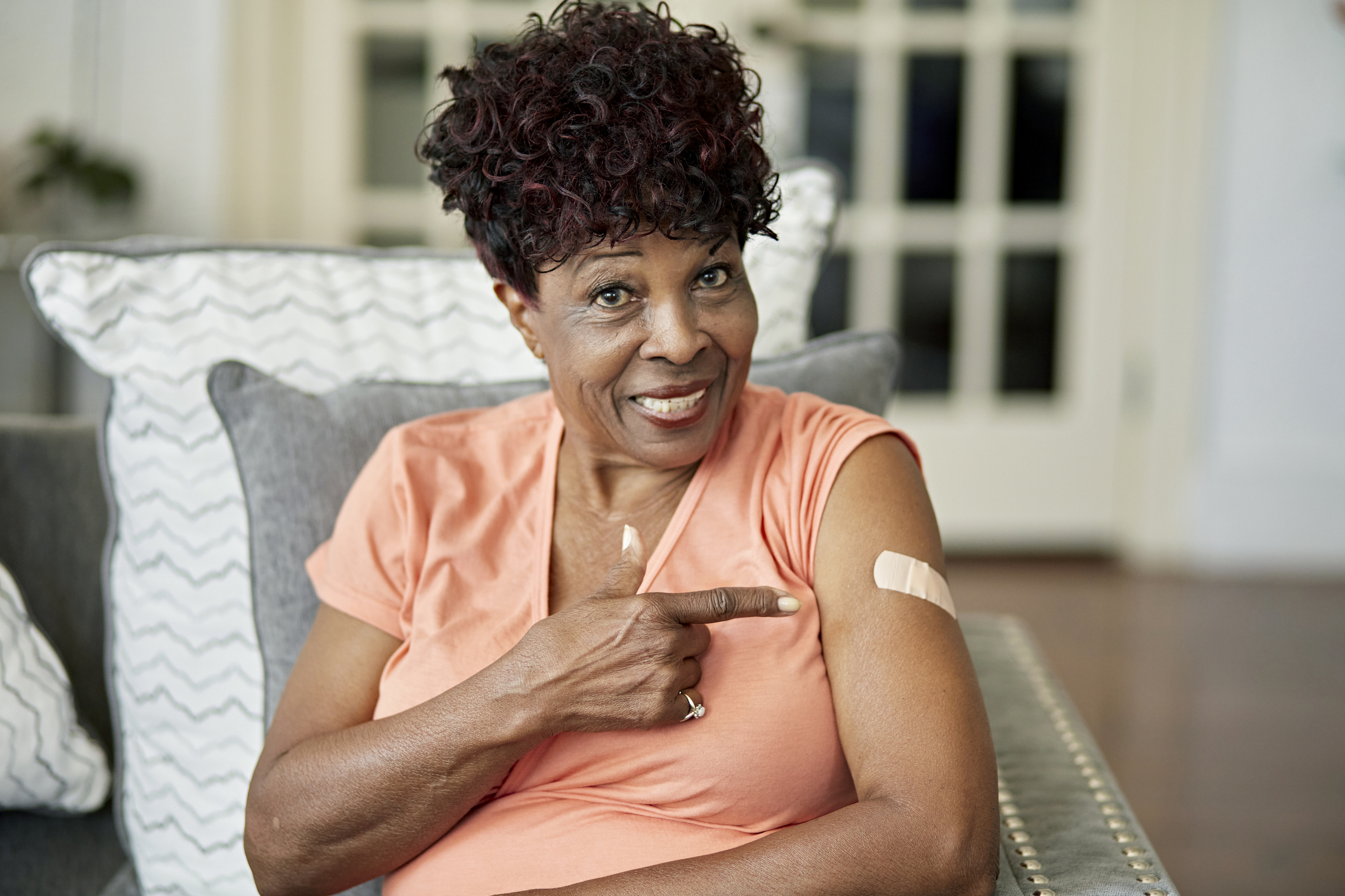 Waist-up view of casually dressed woman sitting in family home and smiling at camera as she gestures with pride at adhesive bandage.
