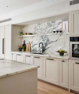 A luxurious kitchen with veined marble splashback and worktops, cream cabinetry, and black accents. A matte black tap and open shelving create a refined look.