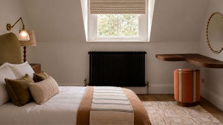 Bedroom with white walls, a window with a Roman blind in the centre, an olive green bed with neutral bedding and a wall sconce above it, a cream rug, and an orange pouffe in the corner.
