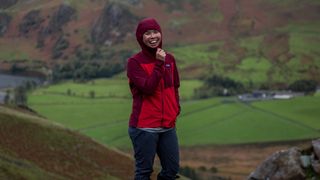 A woman wearing a Patagonia Women’s R1 CrossStrata Hoody with the hood up, standing in grassy hills.