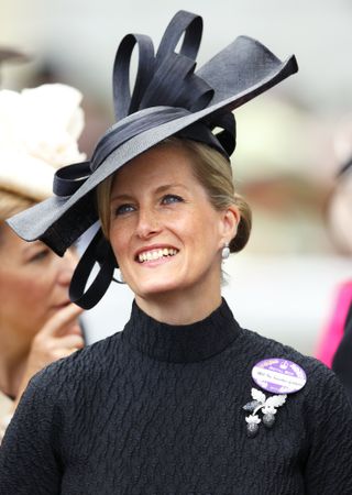 Duchess Sophie wearing a black coat with a black hat at Royal Ascot and smiling