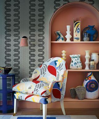 A colourfully covered armchair on a blue rug beside a painted shelf unit of colourful vases.