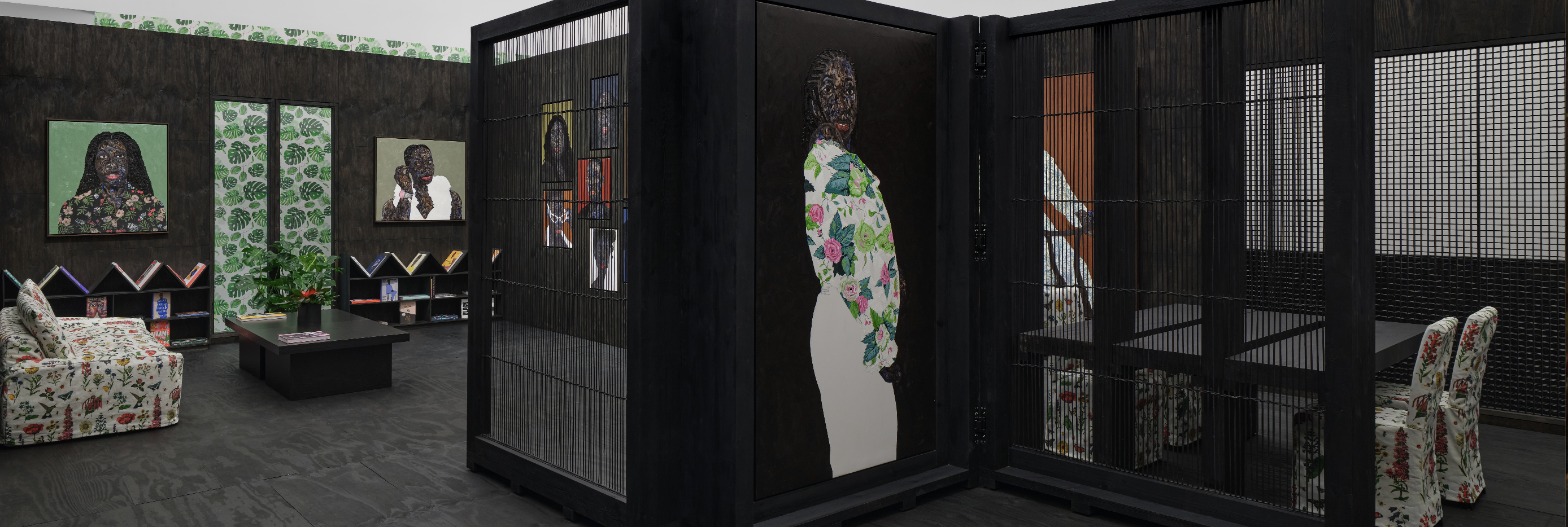 A man wearing a light green floral short-sleeve shirt and matching shorts, a white cap, and sneakers stands in an art gallery with wooden floors and dark teal walls. Three colourful portrait paintings hang behind him, evenly spaced on the wall.