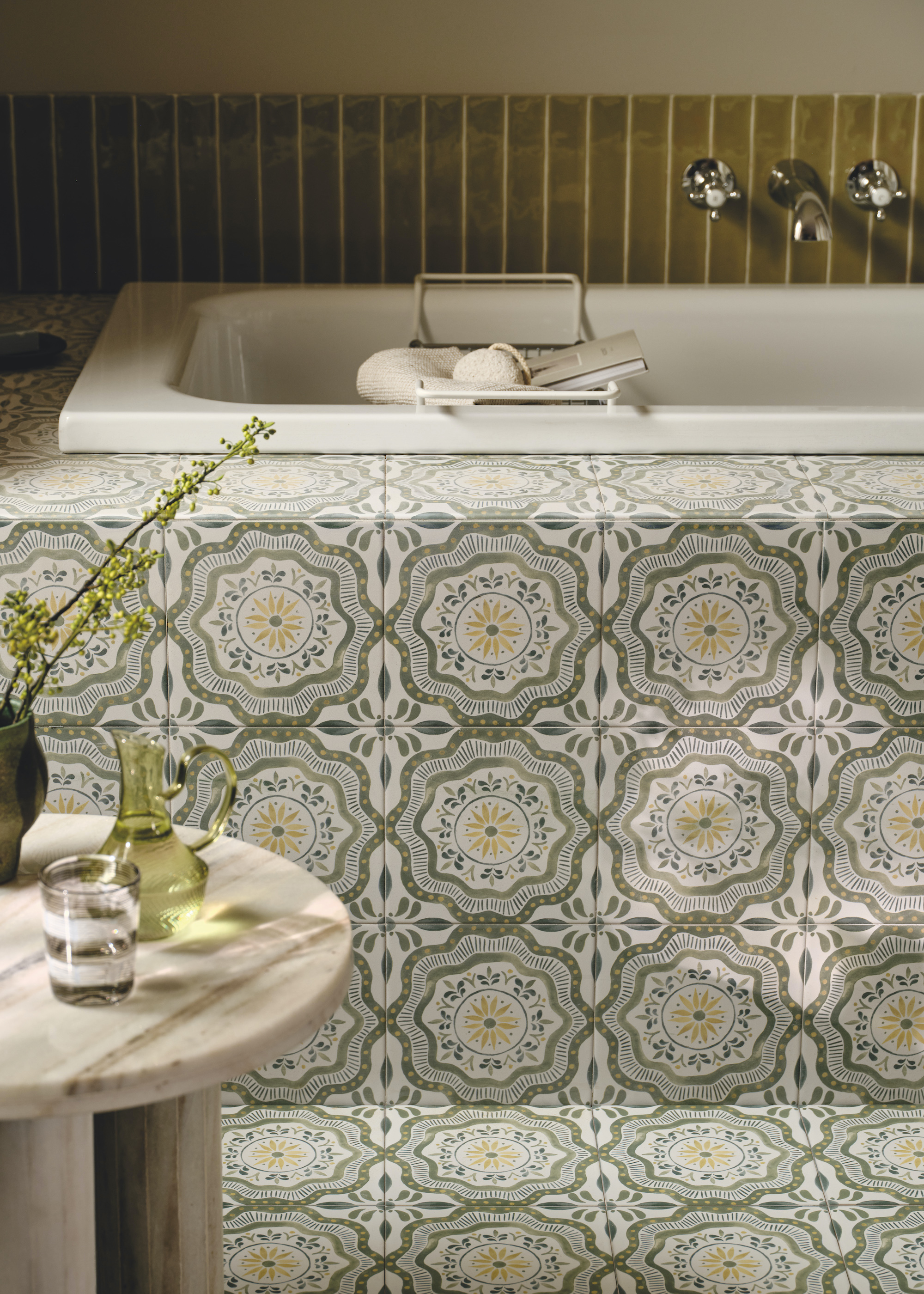 Patterned bath tiled running from the floor of the bathroom to the bathtub itself. There is also a view of a side table with a glass and jug of water