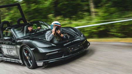 A photographer wearing a helmet leans out of the front trunk of a matte black, modified Porsche convertible, taking a photo while the car speeds down a tree-lined road with motion blur.