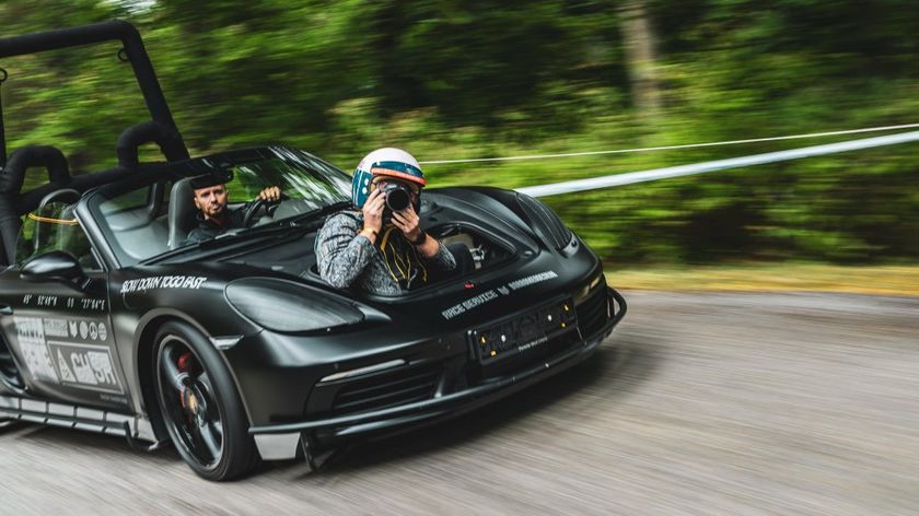 A photographer wearing a helmet leans out of the front trunk of a matte black, modified Porsche convertible, taking a photo while the car speeds down a tree-lined road with motion blur.