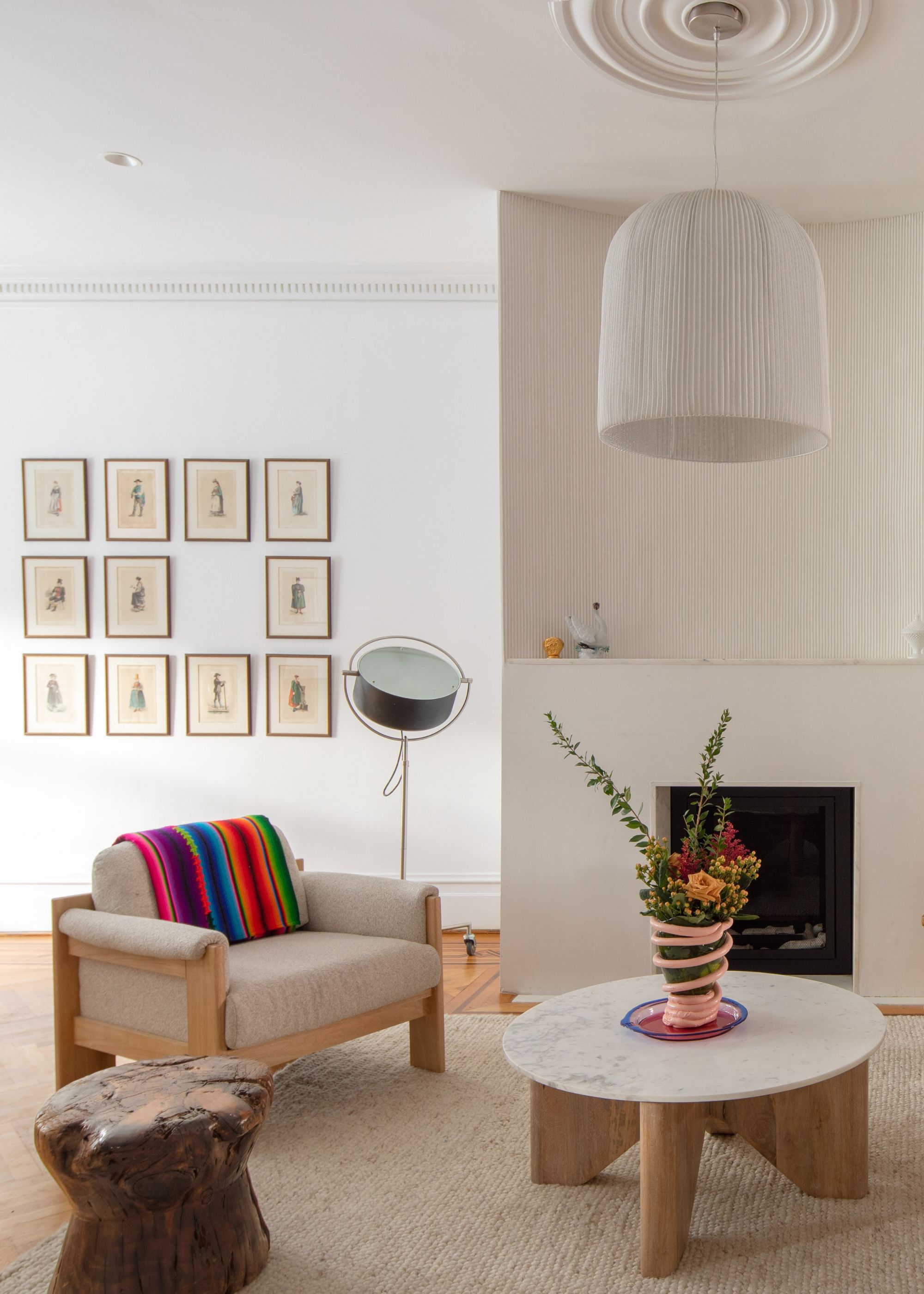 A white living room with a beige armchair, a wood and marble side table, a maximalist vase, a brown stool, a pleated pendant, a wall of frames, a standing lamp, and a vibrant striped throw