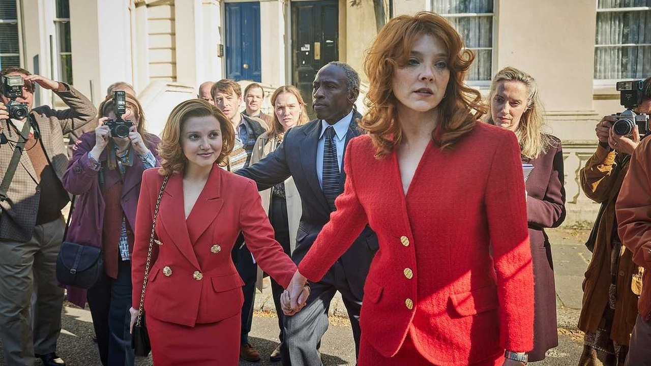 A beaming Jane Andrews (Mia McKenna-Bruce) and glassy-eyed Duchess of York (Natalie Dormer) surrounded by press, hand in hand and wearing matching red suits with gold buttons in a still for ITV's The Lady.