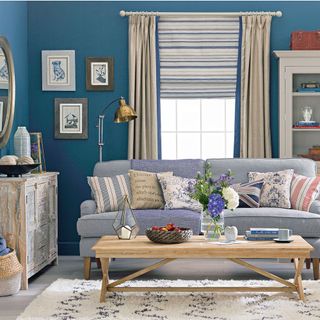 blue living room with cream curtains and blue and cream blinds, a grey sofa with an assortment of cushions, a wooden coffee table and a wooden storage unit