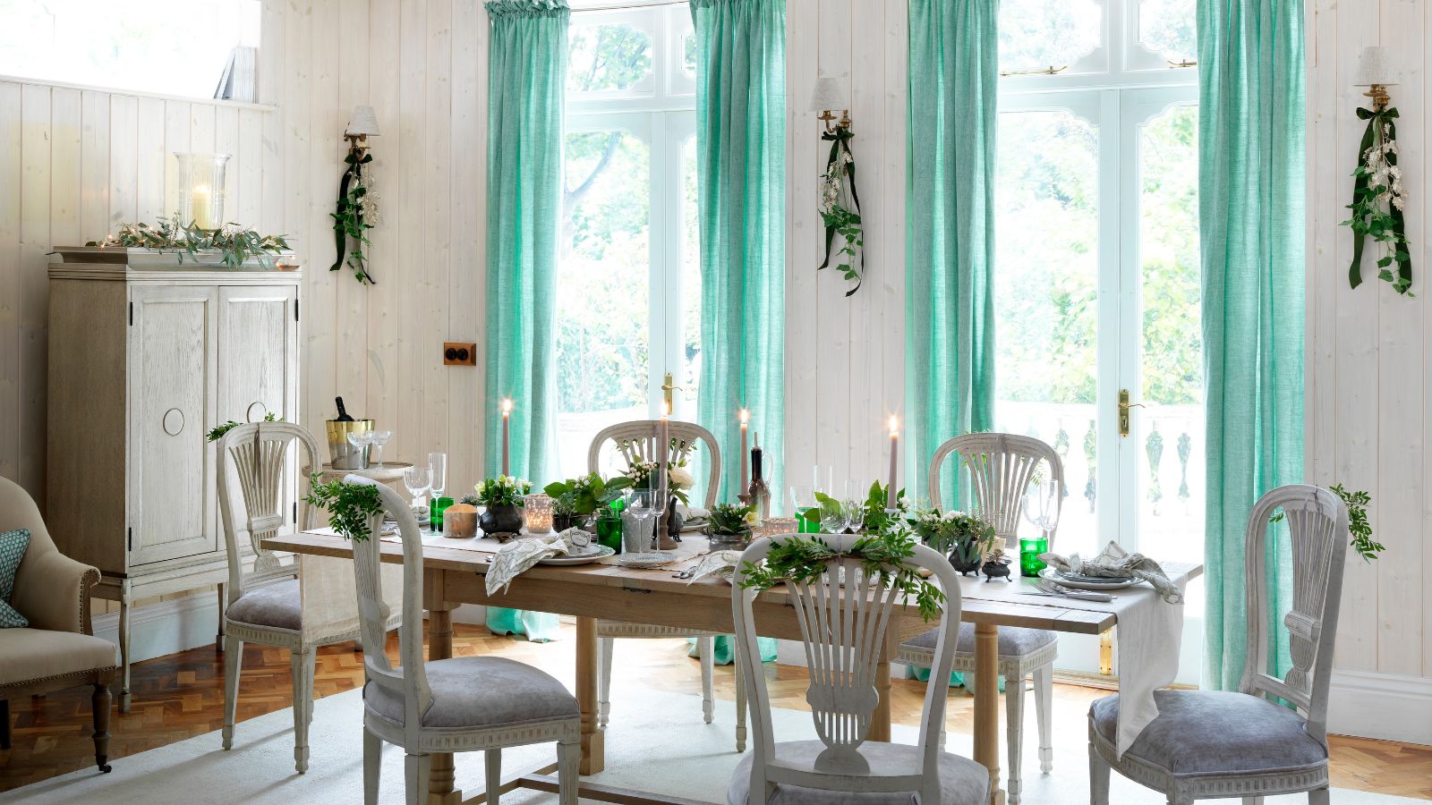 A wooden dining table in a white room set with taper candles. Behind the table are two large windows with blue windows, and around the table are six matching white chairs. To the left is a white cabinet, and beneath the table is a large white rug.
