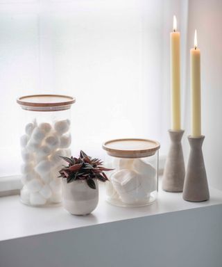 A white bathroom windowsill with two glass jars with cotton balls in them, a plant, and two candle sticks in gray holders