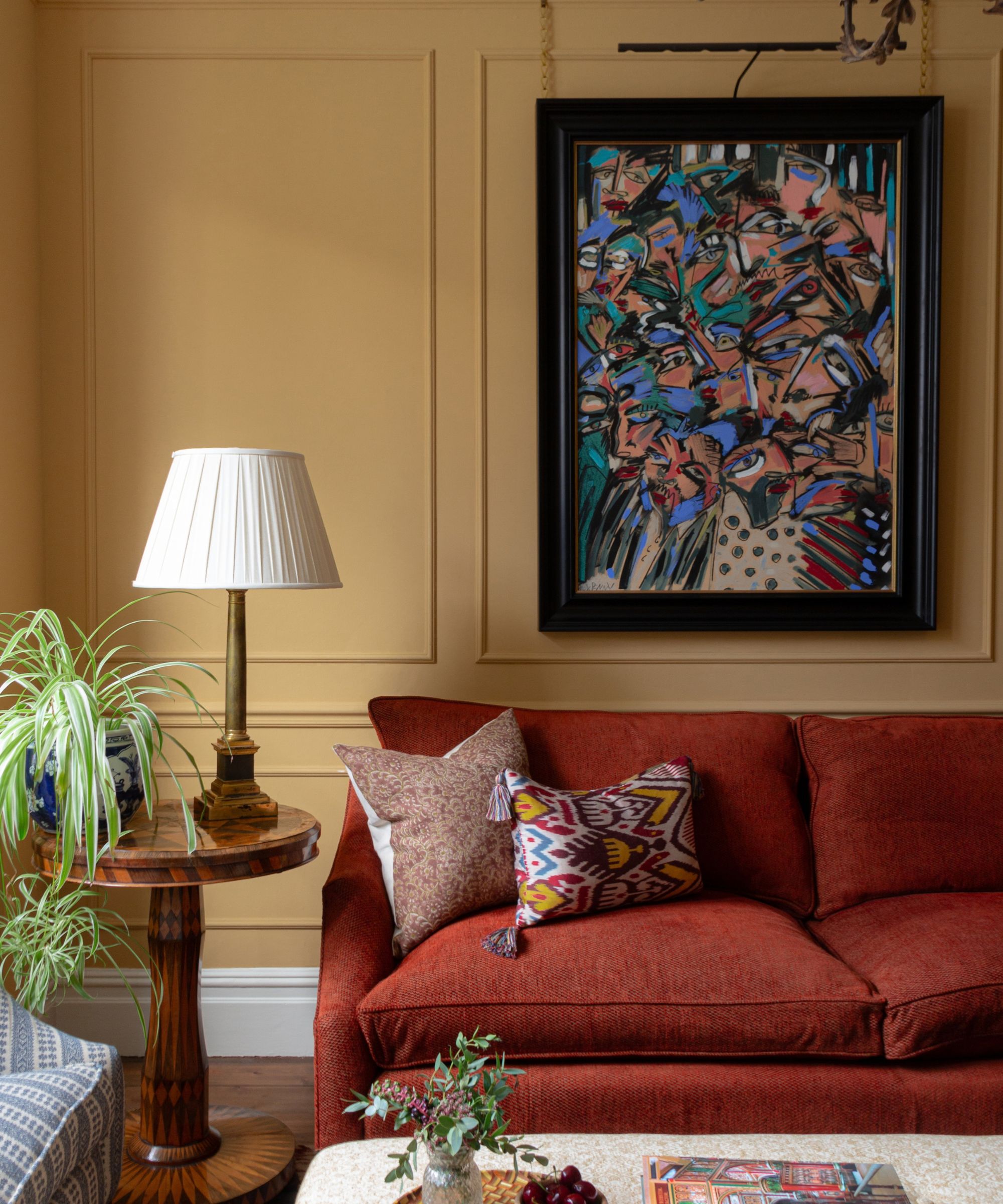Mustard yellow living room with red sofa, a large framed colourful abstract painting on the wall, wooden side table with a pleated table lamp, and an upholstered ottoman with books and vase on top.