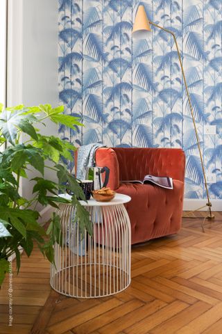 Blue print wallpaper in lounge with herringbone wooden flooring, orange accent chair and white coffee table