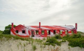 Side view of red painted house surrounded by sand & tall grass