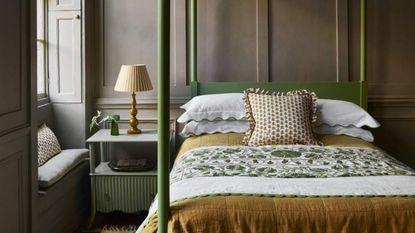 A green four-poster covered in green, gold, and white bedding against brown walls. To the left, a nightstand topped with a lamp.