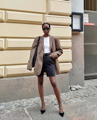 female influencer Sylvie Mus poses on a Paris sidewalk wearing black oval sunglasses, a. tan oversize blazer, white tee, chain necklace, black Bermuda cutoff denim shorts, a black belt, and black suede pumps