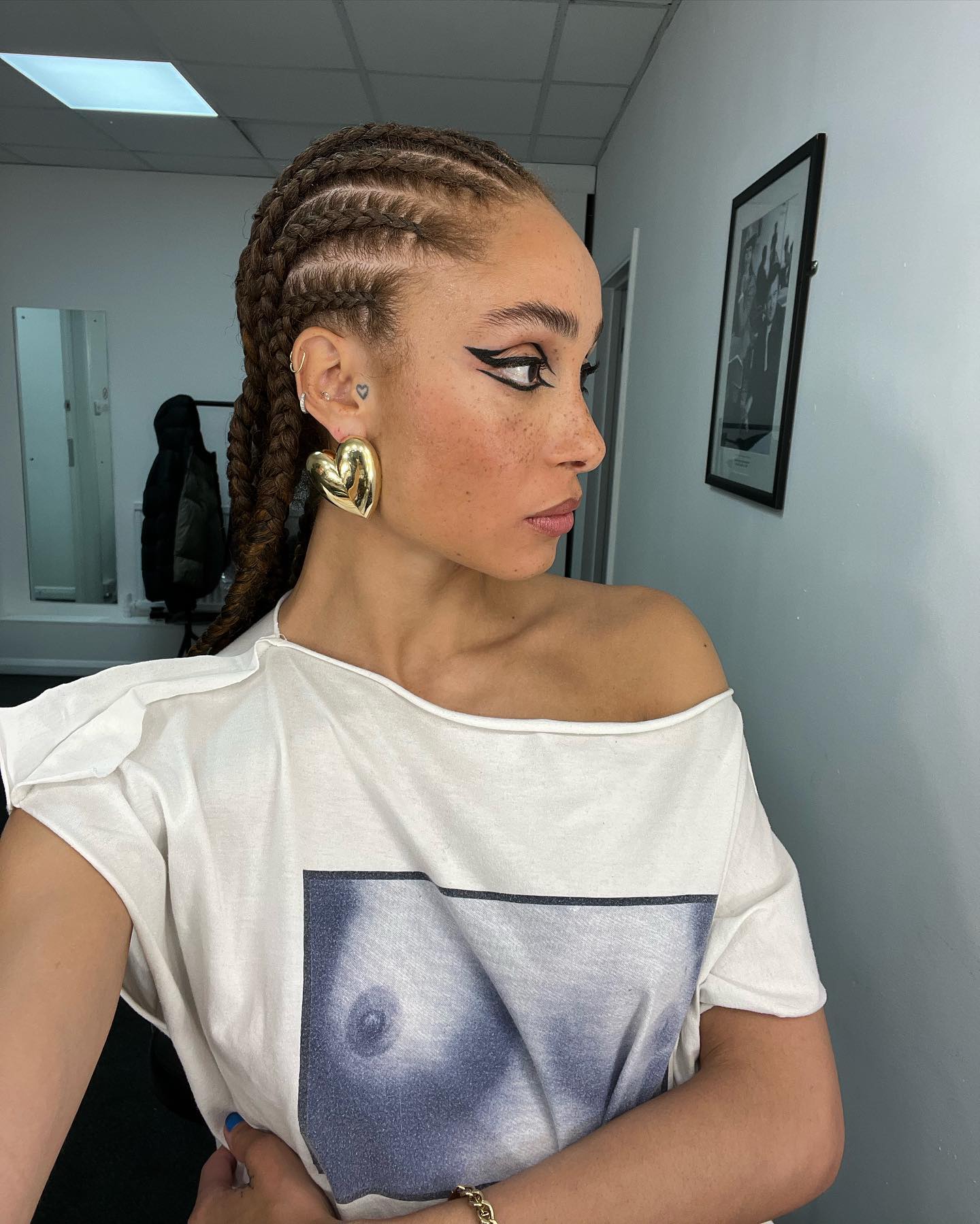 Model Adwoa Aboah with large gold heart earrings, an off the shoulder white top, and stitch braids with graphic black eyeliner