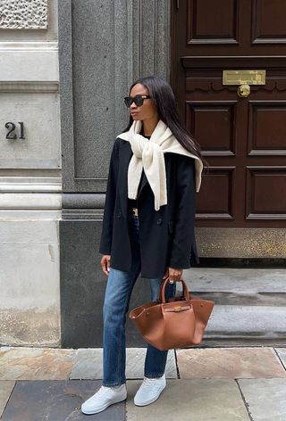 A woman wearing a black oversize blazer with a white cardigan layered over it with skinny jeans, white sneakers, and a brown handbag.