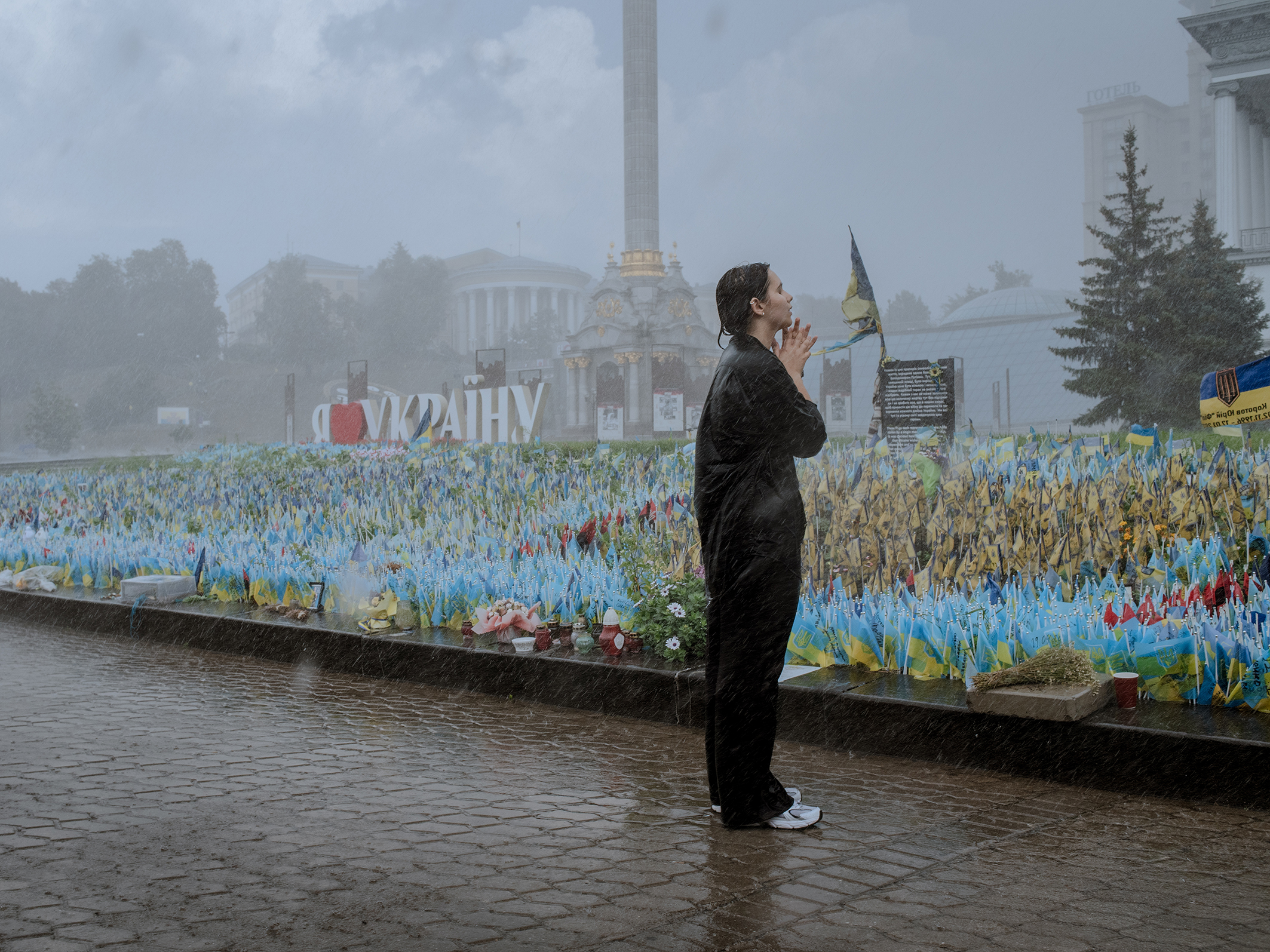 A person prays at a memorial in the rain, surrounded by small blue and yellow flags and flowers. The mood is somber and reflective amidst the downpour