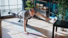 man performing a bird dog exercise on an exercise mat in a living room setting with a window behind him and facing a laptop on a small table.