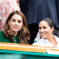 Princess Kate Middleton wears a green dress and has long wavy brown hair and Duchess Meghan Markle wears a white shirt and wears her hair in a ponytail