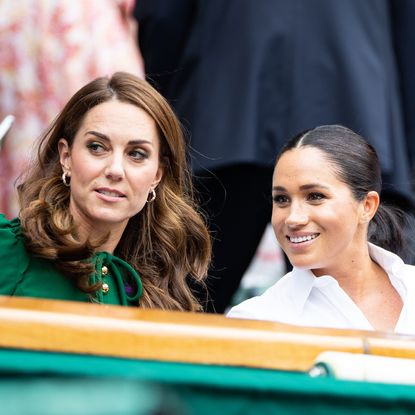 Princess Kate Middleton wears a green dress and has long wavy brown hair and Duchess Meghan Markle wears a white shirt and wears her hair in a ponytail