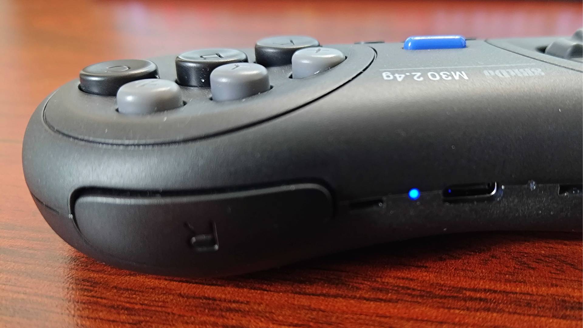 Close up of 8Bitdo M30 controller blue power light next to USB-C port on woodgrain desk.