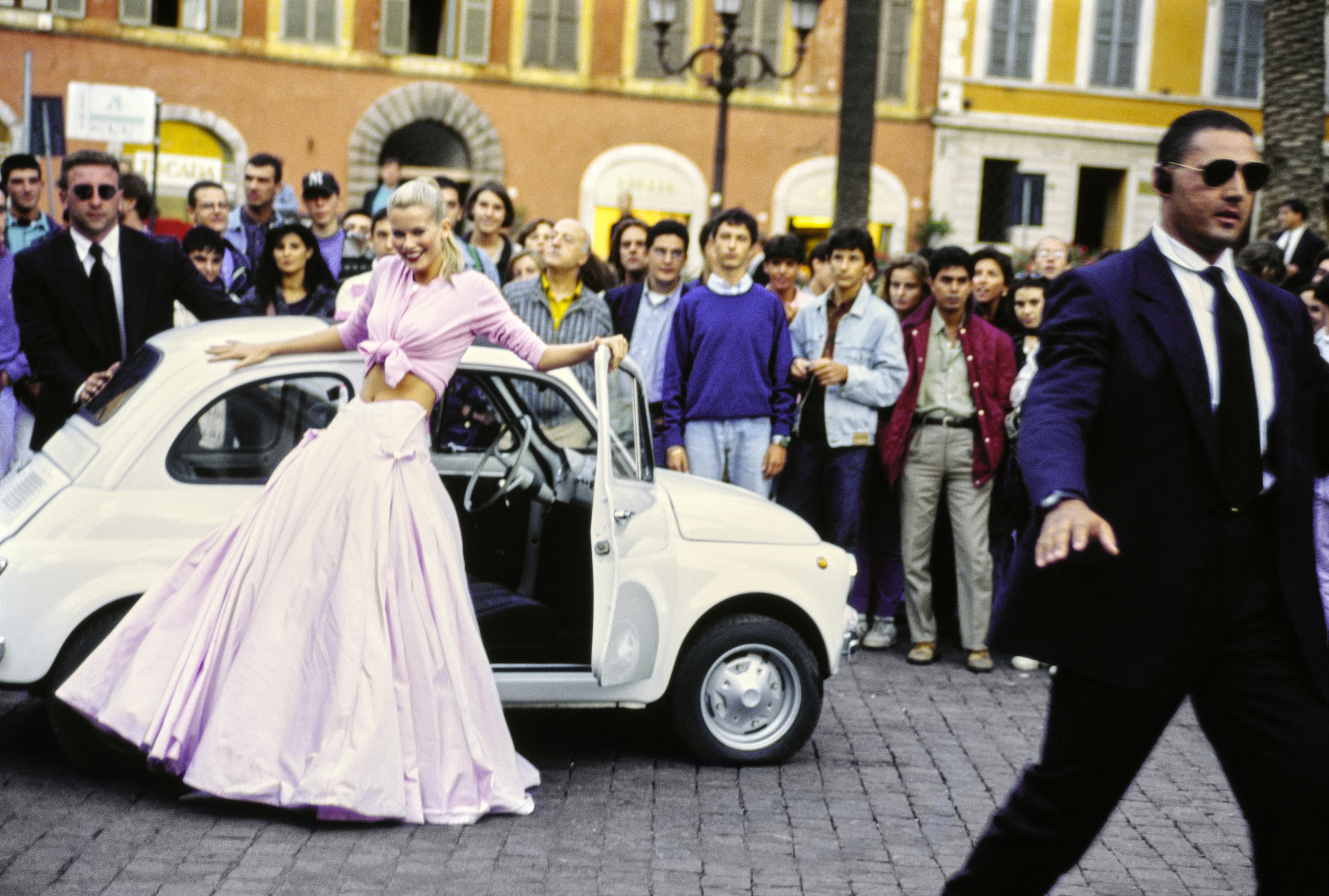 Model Claudia Schiffer leans against a 1967 white Fiat 500 on the street in Rome, Italy. She is wearing a waist-tied pink wool cardigan by Genny over a Taroni silk taffeta skirt by Atelier Versace.