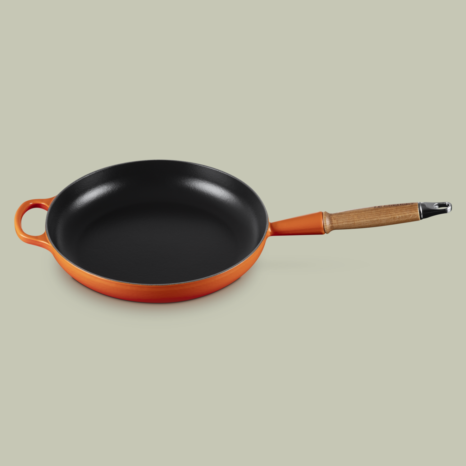 An orange cast iron frying pan with a wooden handle from Le Creuset, against a soft gray green background