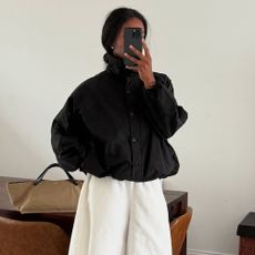 Influencer @nlmarilyn takes a mirror selfie indoors, wearing a black windbreaker jacket, fastened to the top with a white skirt and black heels.
